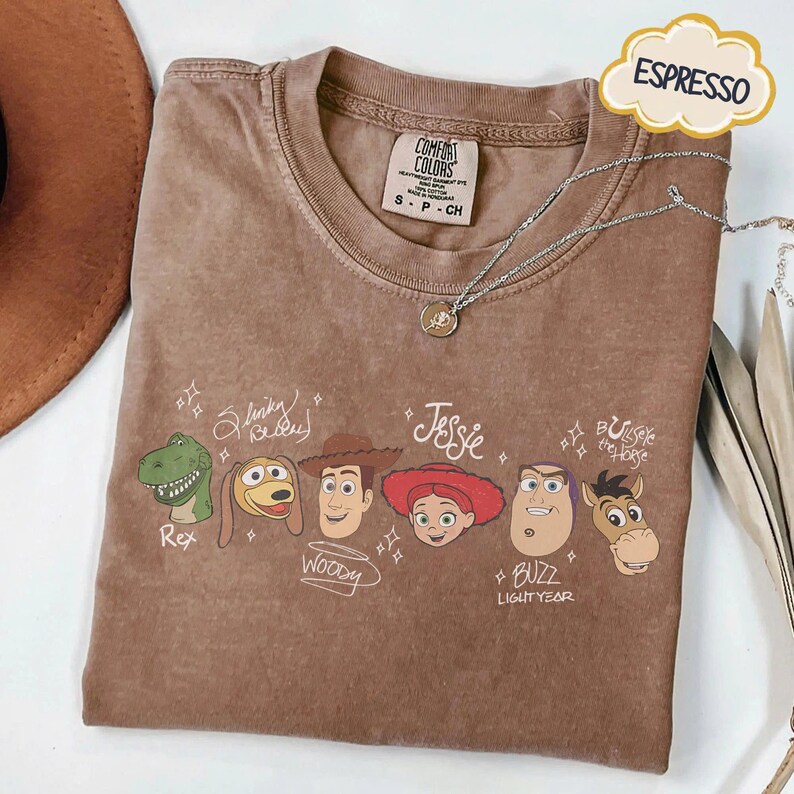 May include: An espresso-colored t-shirt featuring a graphic print of six animated characters with their names written in cursive script: Rex, Slinky Dog, Woody, Jessie, Buzz Lightyear, and Bullseye the Horse, accented with small star symbols.