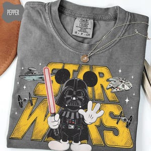 Puede incluir: Camiseta gris con un dise&ntilde;o de Star Wars, con Darth Vader al estilo Mickey Mouse, sosteniendo un sable de luz rojo. El texto "STAR WARS" es amarillo, con naves espaciales y estrellas. La camiseta tiene una etiqueta Comfort Colors.