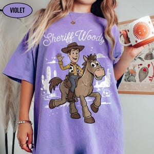 May include: A violet t-shirt featuring a graphic print of Sheriff Woody riding a horse. The text "Sheriff Woody" is printed in white script above the characters. The design includes a desert scene with cacti and a skull. The shirt has a crew neckline.