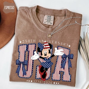 Może przedstawiać: Koszulka w kolorze espresso z grafiką Myszki Minnie w patriotycznej sukience i kokardzie. Projekt zawiera napis 250TH ANNIVERSARY, USA oraz 1776-2026, podkreślony czerwono-biało-niebieskimi literami w paski oraz niebieskimi kokardami w kropki.