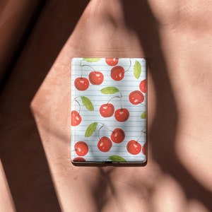 May include: A white and blue striped case with a red cherry pattern. The case is for an electronic device like a tablet or e-reader.