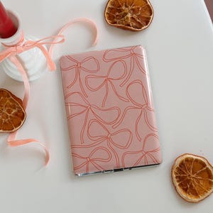 Pode incluir: Um espelho compacto rosa com uma moldura prateada e um padrão de laços vermelhos na frente.