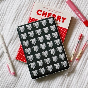 May include: A black and white patterned case with a design of silver hearts. The case is sitting on a white blanket with a red and white book titled "CHERRY" and a pink highlighter with the text "TOWER 28" on it.