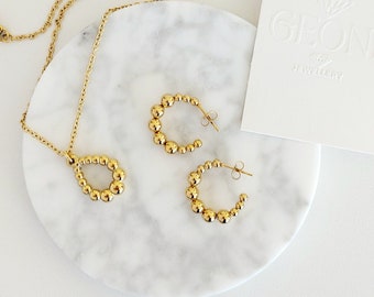 Op de afbeelding: Een gouden ketting- en oorstekerset met een traanvormige hanger en creolen. De sieraden zijn gemaakt van kleine, ronde kralen.