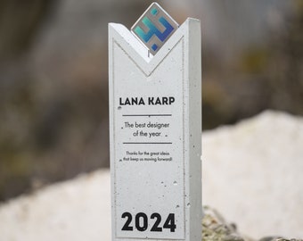 Puede incluir: Un premio de hormigón gris con un logotipo blanco y azul en la parte superior. El premio dice "LANA KARP El mejor diseñador del año Gracias por las grandes ideas que nos mantienen avanzando! 2024"