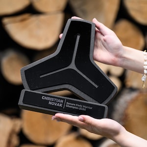 May include: A black, three-pointed trophy with a matte finish and a rectangular base. The base features white text that reads: CHRISTIAN NOVAK Secure Code Warrior Champion 2026. The trophy has a distinct geometric shape with a central Y-shaped cutout.