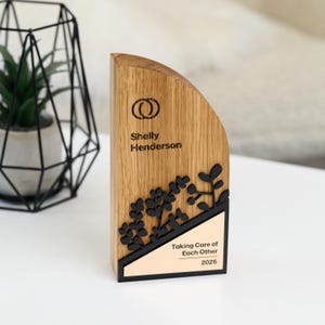 May include: A wooden award plaque with a black and gold design. The plaque features a stylized plant design, the text "Taking Care of Each Other", and the year "2025". The name "Shelly Henderson" is engraved on the plaque.