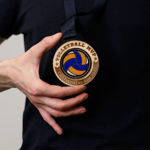 Peut inclure: Une médaille de MVP de volleyball en bois avec un graphique de volleyball bleu et jaune. La médaille indique "Volleyball MVP Continental Cup 2025".