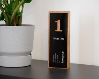 May include: A wooden award plaque with a black centre and the number "1" in a light wood tone. The plaque includes the name "John Doe" and the text "ideagroup". A potted plant sits to the left.