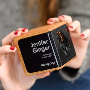 Può includere: Un cubo di legno con una placca nera. La placca riporta il testo "Jenifer Ginger" e "Miglior Manager dell'anno 2023" e il logo "ideagroup". Il cubo ha un design a forma di impronta digitale su un lato.