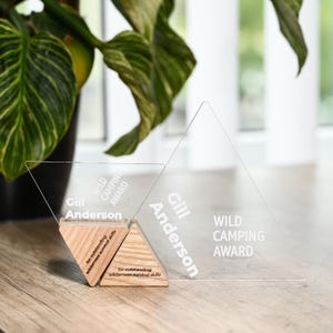 May include: A clear acrylic award in the shape of a mountain with the text "WILD CAMPING AWARD" and "Gill Anderson" engraved on it. The award is on a wooden base.