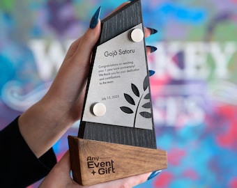 May include: A black and brown wooden award plaque with a silver metal plate. The plate has the text "Gojo Satoru" and a congratulatory message for reaching a work anniversary. The date "July 15, 2023" is also included. The plaque has a leaf design and the text "Any Event + Gift" is on the bottom.