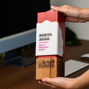 Peut inclure: Un trophée rectangulaire composé d'une partie supérieure en bois teinté rouge avec une plaque en acrylique blanc portant le texte MARIYA JIVAJI et Arts in education award, fixé sur une base en bois clair naturel gravée Arts Awards.