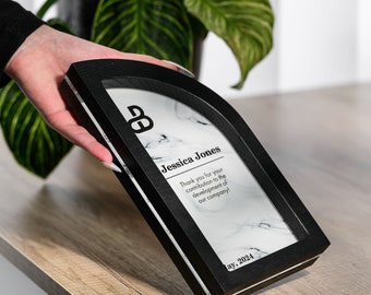 Puede incluir: Una placa de madera negra con un diseño de mármol y un inserto de acrílico transparente. La placa tiene una forma de arco redondeado y presenta el texto "B", "Jessica Jones", "¡Gracias por su contribución al desarrollo de nuestra empresa!" y "ay, 2024".