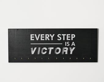May include: Black wooden sign with the text "EVERY STEP IS A VICTORY" in white lettering. The sign has small gold hooks along the bottom.