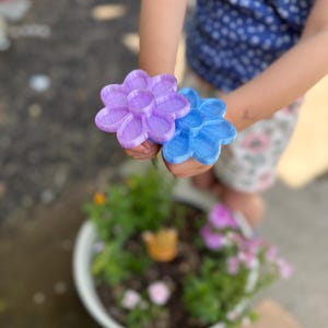 May include: A pair of 3D-printed flower-shaped toys, one lavender and one blue, held in a child's hands. The flowers have a textured surface and a detailed petal design. The background includes a flower pot with greenery and other flowers.