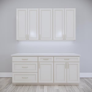 Pode incluir: Armários de cozinha creme com ferragens douradas. Os armários superiores são montados acima de uma bancada com armários inferiores e gavetas. Os armários têm um design clássico e estão colocados contra uma parede cinza claro.