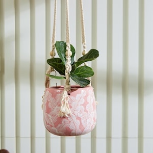 May include: A pink ceramic planter with a white fern leaf pattern suspended by a cream-colored macrame cord featuring wooden beads. A small green fiddle leaf fig plant is potted inside the rounded vessel, which hangs from a central point.