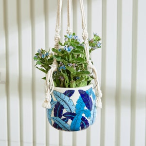 May include: A hanging planter with a white ceramic pot featuring a blue leaf design. The pot holds green plants with small blue flowers. The planter is suspended by a cream-colored macrame hanger, creating a decorative and natural aesthetic.