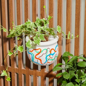 May include: A white ceramic planter with a colorful abstract design featuring blue, green, orange, and brown lines. The planter is filled with a green leafy plant with variegated leaves.