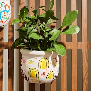 May include: A white ceramic planter with a hand-painted design featuring yellow, red, and brown accents. The planter is filled with green leafy plants and is hanging on a wooden slat wall.