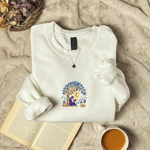 May include: A white crewneck sweatshirt with an embroidered design of a person reading under a tree with a crescent moon and stars. A silver necklace with a dark blue pendant is visible. The sweatshirt is on a soft, textured surface.