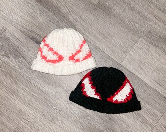 May include: Two knitted beanies with eye designs. One beanie is white with red eyes, and the other is black with white and red eyes.