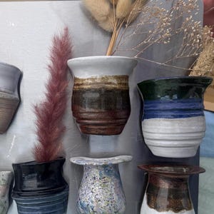 Puede incluir: Un tablero de exhibición rectangular de metal gris con nueve pequeños recipientes de cerámica montados en la pared en varios esmaltes, incluyendo verde moteado, marrón, azul y blanco. Varios tallos botánicos secos están colocados en las tazas.