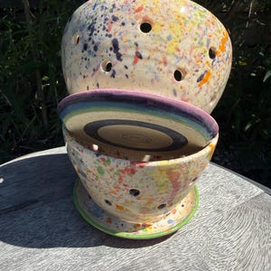 May include: A ceramic berry bowl set featuring a cream-colored glaze with a multi-colored paint splatter pattern. The set includes a colander bowl with drainage holes, a matching saucer, and a flat circular lid with a rainbow-colored rim.