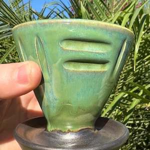 May include: A green ceramic pour-over coffee dripper with a black ceramic base. The dripper has a unique design with three horizontal ridges and a textured surface.