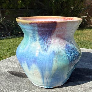 Puede incluir: Un jarrón de cerámica con un borde ancho y acampanado y un cuerpo redondeado. La superficie presenta un esmalte brillante y multicolor con patrones fluidos en forma de goteo en tonos azul cielo, índigo profundo, turquesa y toques de melocotón y crema.