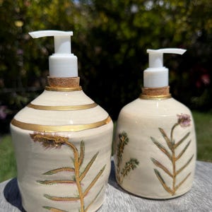 Puede incluir: Dos dispensadores de jabón de cerámica con bombas blancas. Los dispensadores son de color blanquecino con detalles dorados y diseños botánicos en verde y dorado. Uno tiene una banda dorada horizontal, y ambos tienen bases de corcho.