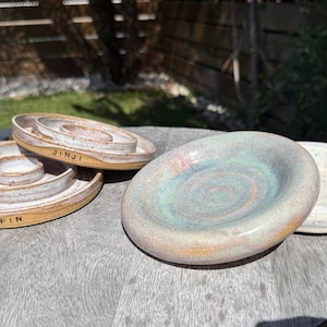 May include: Four circular ceramic dishes with raised edges. Two dishes feature a central raised ring and the stamped text JINJI and FIN on the outer rim. The other two dishes have a smooth, concave surface with a speckled glaze in blue, green, and white.