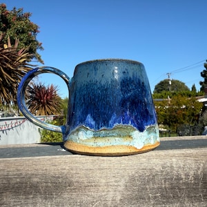 Könnte beinhalten: Eine handgefertigte Keramiktasse mit blauer Glasur, die einen Farbverlauf von dunkel- zu hellblauen Farbtönen aufweist. Die Tasse hat einen breiten, stabilen Henkel und eine strukturierte Oberfläche. Der Boden hat eine beige Farbe. Die Tasse steht auf einer Holzoberfläche.
