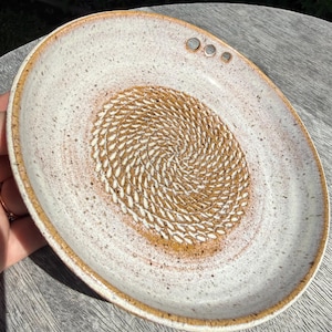 Può includere: Un piatto grattugia in ceramica rotondo con smalto bianco sporco maculato e bordo marrone. Il centro presenta una texture in rilievo con un motivo a spirale color ocra per grattugiare il cibo. Tre piccoli fori circolari sono posizionati vicino al bordo.