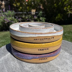 May include: A stack of handmade ceramic bowls in varying colors, including white, yellow, and purple. Each bowl has a unique glaze and is inscribed with a word: MARSHMALLOW, YAMPER, and TONKS. The bowls are stacked on a wooden surface.