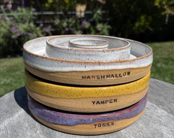 May include: A stack of three round ceramic bowls with a speckled finish. The top bowl is white, the middle is yellow, and the bottom is purple. Each bowl features a stamped name on the side: MARSHMALLOW on the white, YAMPER on the yellow, and TONKS on the purple.