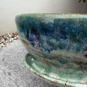Peut inclure: Un bol de plantation en céramique avec sa soucoupe assortie, présentant un émail réactif dans des tons de bleu profond, de sarcelle et de vert forêt. La surface a une texture mouchetée avec des coulures visibles et des variations de couleur.