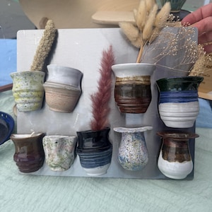 May include: A collection of small, handcrafted ceramic vases in various colors and patterns. The vases are displayed on a gray rectangular board, with dried flowers and grasses adding a natural touch. The vases feature a variety of shapes and finishes.