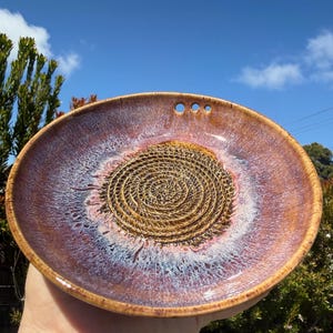 Puede incluir: Un plato de cerámica hecho a mano con un diseño en espiral en el centro. El plato tiene un borde marrón y un interior vidriado morado y blanco. Tres pequeños agujeros están cerca del borde. El plato se sostiene contra un cielo azul.