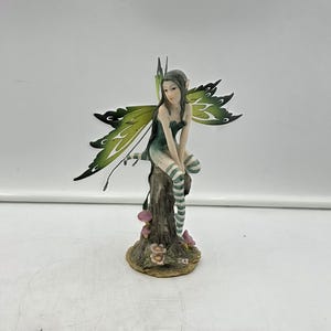 May include: A figurine of a fairy with dark hair and green wings, wearing a dark top and striped stockings, sitting on a brown tree stump base decorated with small pink flowers. The fairy has a slender build and is positioned in a seated pose.