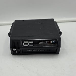 May include: A black Craig eight-track stereo player featuring a rectangular housing with a front-facing control panel. The unit includes two silver push buttons, a volume slider, and a small display area with the brand name Craig printed in white.