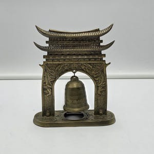 May include: A bronze-colored metal decorative structure shaped like a traditional gate with a curved roof and intricate relief carvings. A small, matching metal bell hangs from the center of the archway, suspended by a thin metal link.