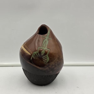May include: A small, asymmetrical brown ceramic vase with a dark, textured base. The upper portion features a smooth, polished finish with a carved, light green stylized figure etched into the surface, and a single small opening at the top.