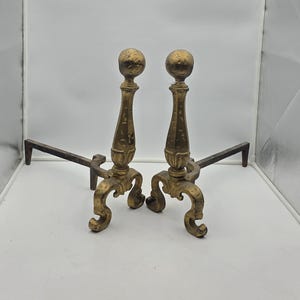 May include: A pair of matching brass fireplace andirons featuring a vertical post topped with a spherical finial, supported by a curved, ornate base with scrollwork details, and extending into a long, straight horizontal iron support bar.