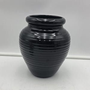 Può includere: Un vaso in ceramica nero lucido con corpo arrotondato e bordo largo e svasato. La superficie esterna presenta una serie di scanalature orizzontali e concentriche che avvolgono l'intera circonferenza del recipiente, dalla base al collo.