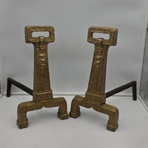 May include: A pair of gold-colored metal fireplace andirons featuring a textured, tapered vertical post with a rectangular handle at the top and a wide, arched base. Each andiron includes a long, dark, L-shaped iron support bar extending backward.