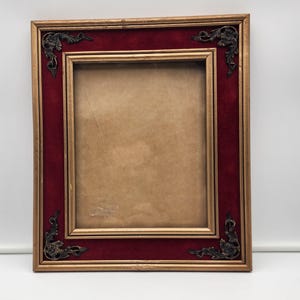 Puede incluir: Un marco de fotos rectangular con un borde dorado y un borde interior de terciopelo rojo oscuro. Las esquinas están adornadas con elementos decorativos de metal oscuro y ornamentado. El marco está diseñado para contener una fotografía o una obra de arte.
