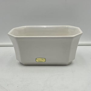 May include: A white ceramic rectangular planter with beveled corners and a smooth finish. A small rectangular gold-colored sticker with black text is centered on the front face of the vessel. The planter has a wide opening and a flat base.