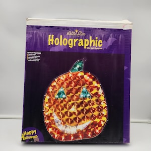 May include: A holographic pumpkin-shaped mini light sculpture in a box. The pumpkin has a smiling face with green triangle eyes and a green stem. The box is purple with the words "Holographic" and "Happy Halloween".